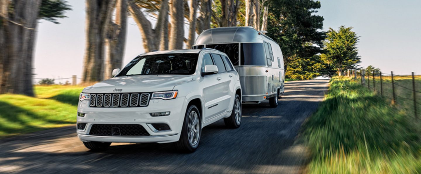 A 2020 Jeep Grand Cherokee towing a trailer on a single-lane road.