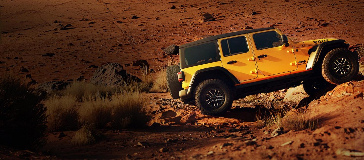 A passenger-side profile of a gold 2026 Jeep Wrangler Willys 392 crawling up a steep, rocky trail in the desert